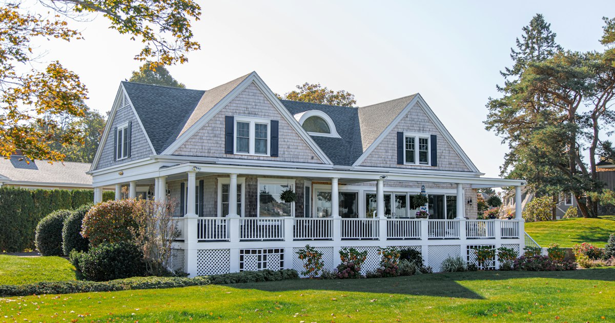 A grey shingled house with a white wraparound porch and dormer windows, seen across a mown front lawn