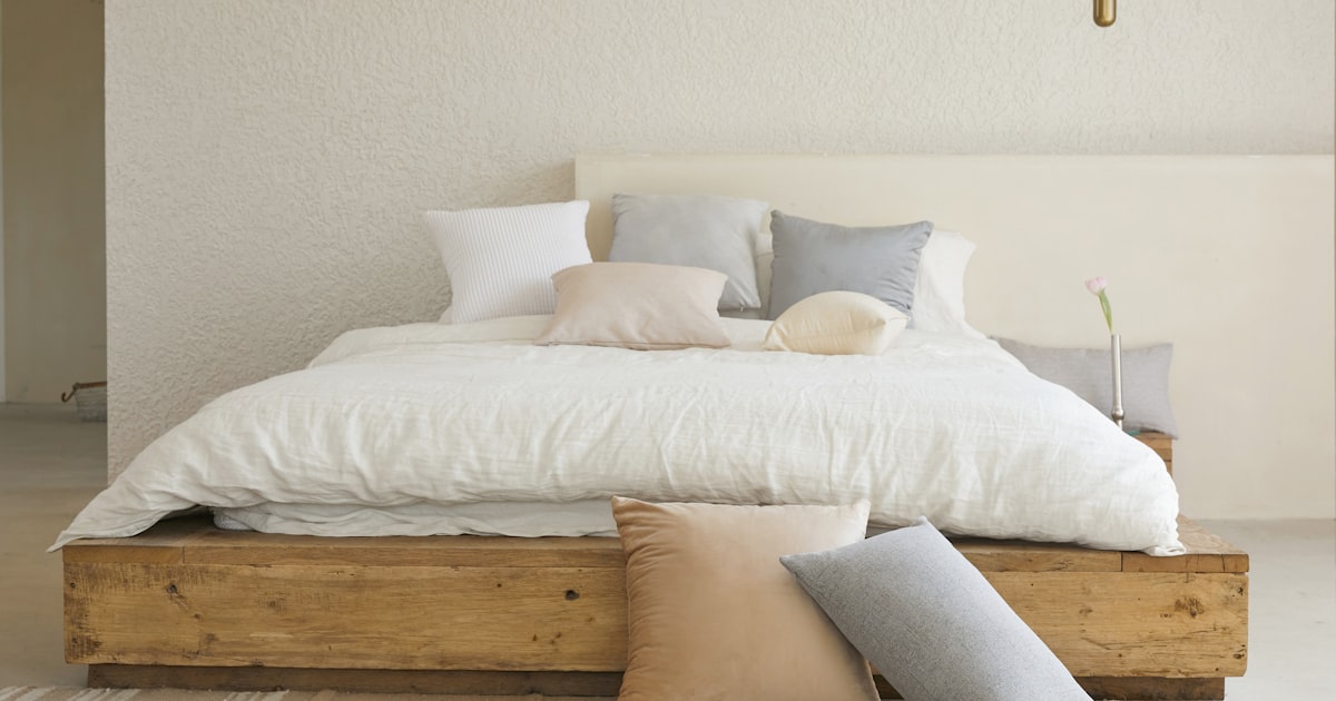 A low wooden platform bed with white bedding against a plain neutral wall