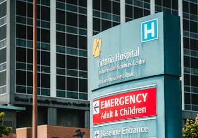 A teal hospital wayfinding sign reading Victoria Hospital, with a red Emergency arrow board beneath it and a tall glass hospital block rising behind